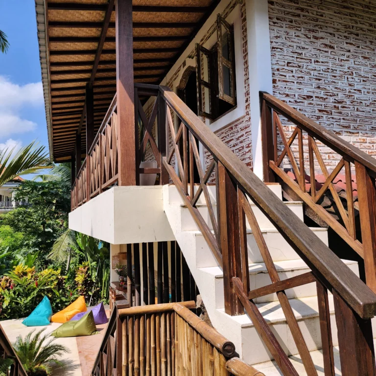 Tropical outdoor area with wooden staircase, brick wall, colorful bean bags, and lush greenery.
