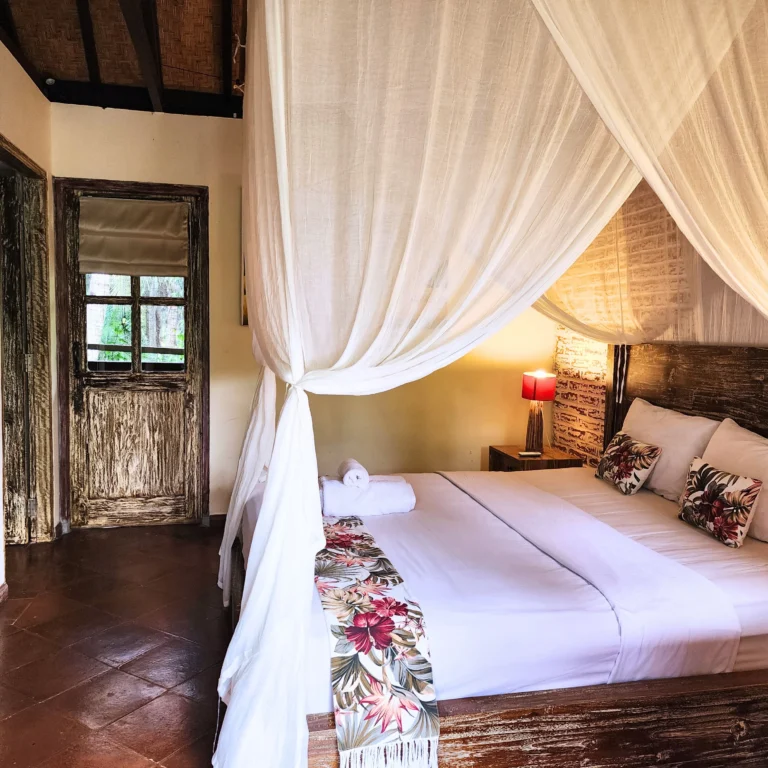 Rustic Balinese bedroom with white canopy bed, floral accents, wooden furniture, and warm ambient lighting.