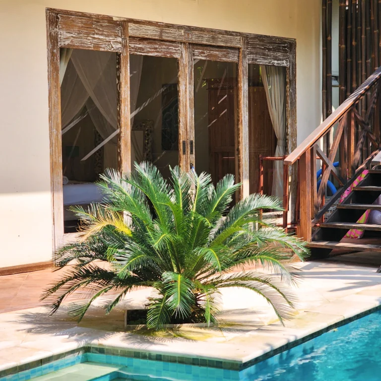 Poolside view with tropical plants, wooden structure, sliding glass doors, and a bedroom with mosquito net.