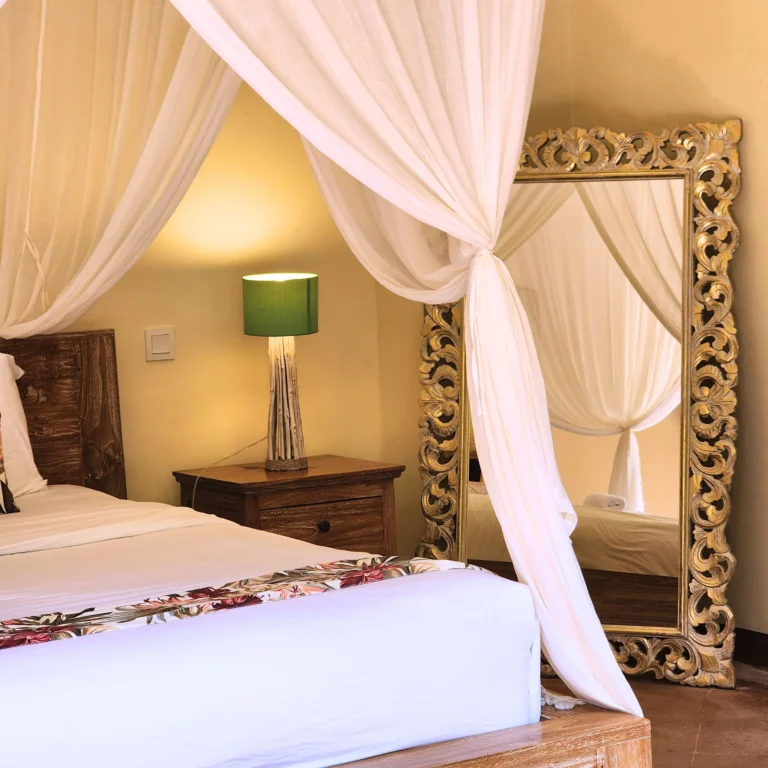 Elegant Balinese-style bedroom with white canopy bed, floral runner, wooden nightstand, and ornate gold-framed mirror.