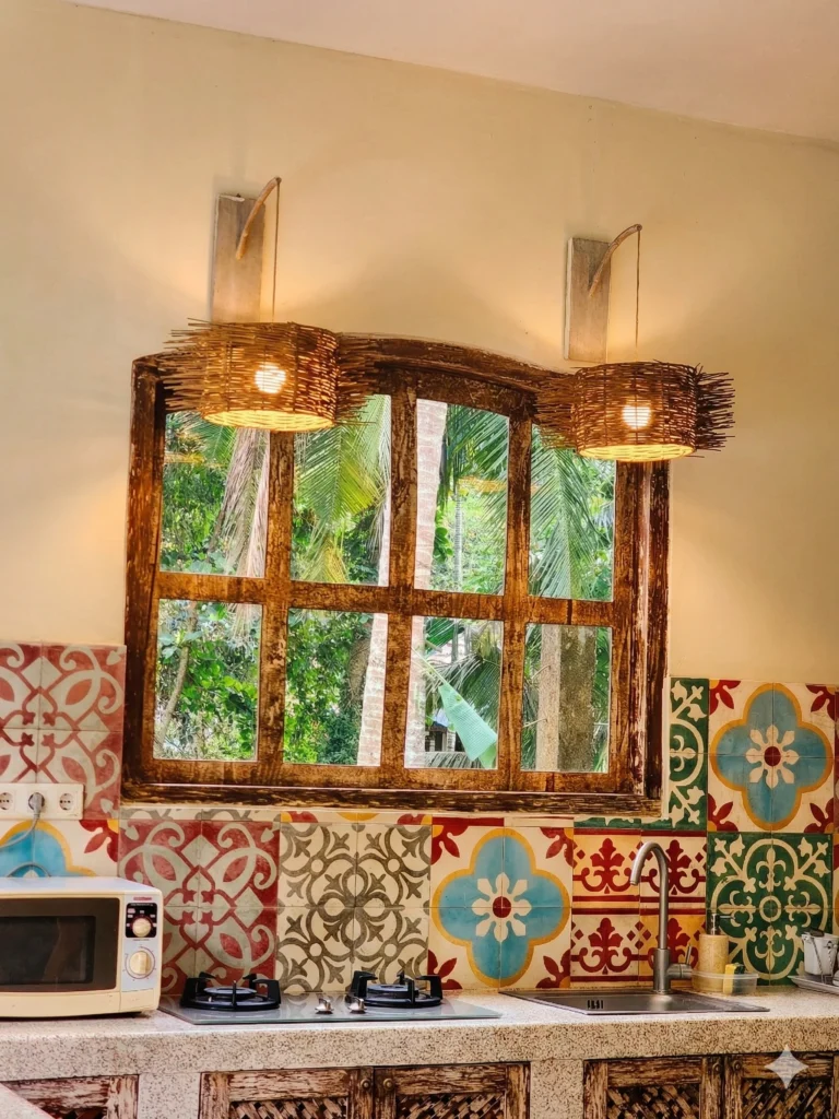 Rustic kitchen with patterned tile backsplash, wooden window, woven wall lights, and tropical greenery outside.